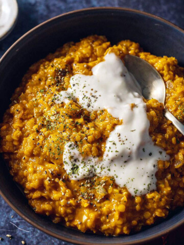 A bowl filled with red lentil dhal with a spoon on the side and coconut yogurt on top.