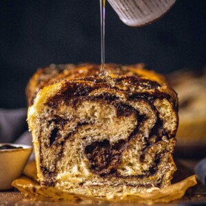 Syrup being drizzled on a cut open chocolate babka showcasing soft dough with swirled layers of chocolate.