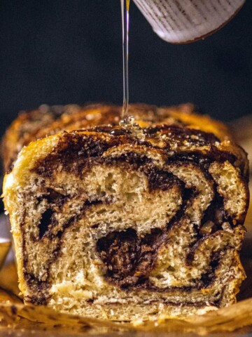 Syrup being drizzled on a cut open chocolate babka showcasing soft dough with swirled layers of chocolate.