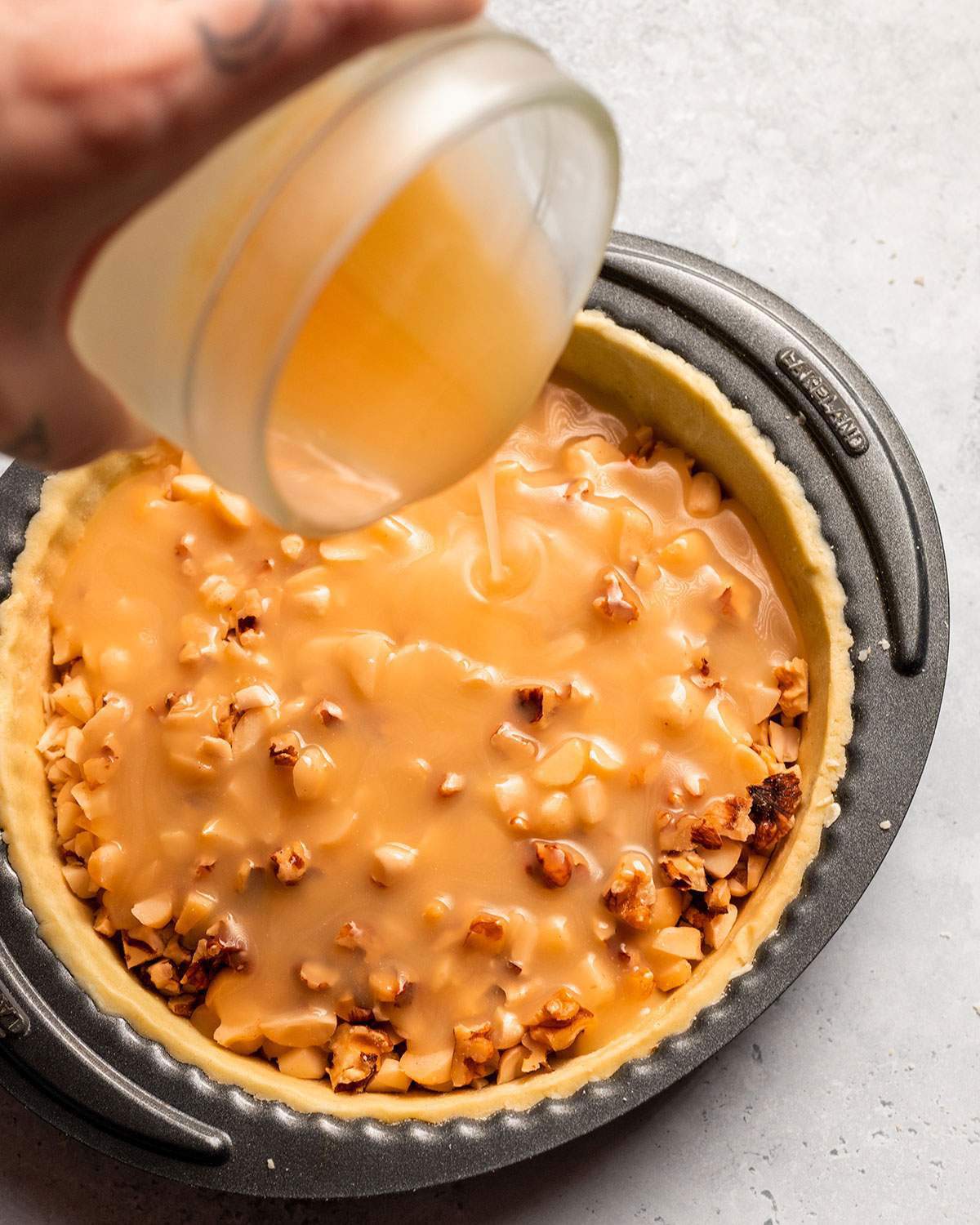 Pouring caramel sauce onto nuts in a vegan tart base.