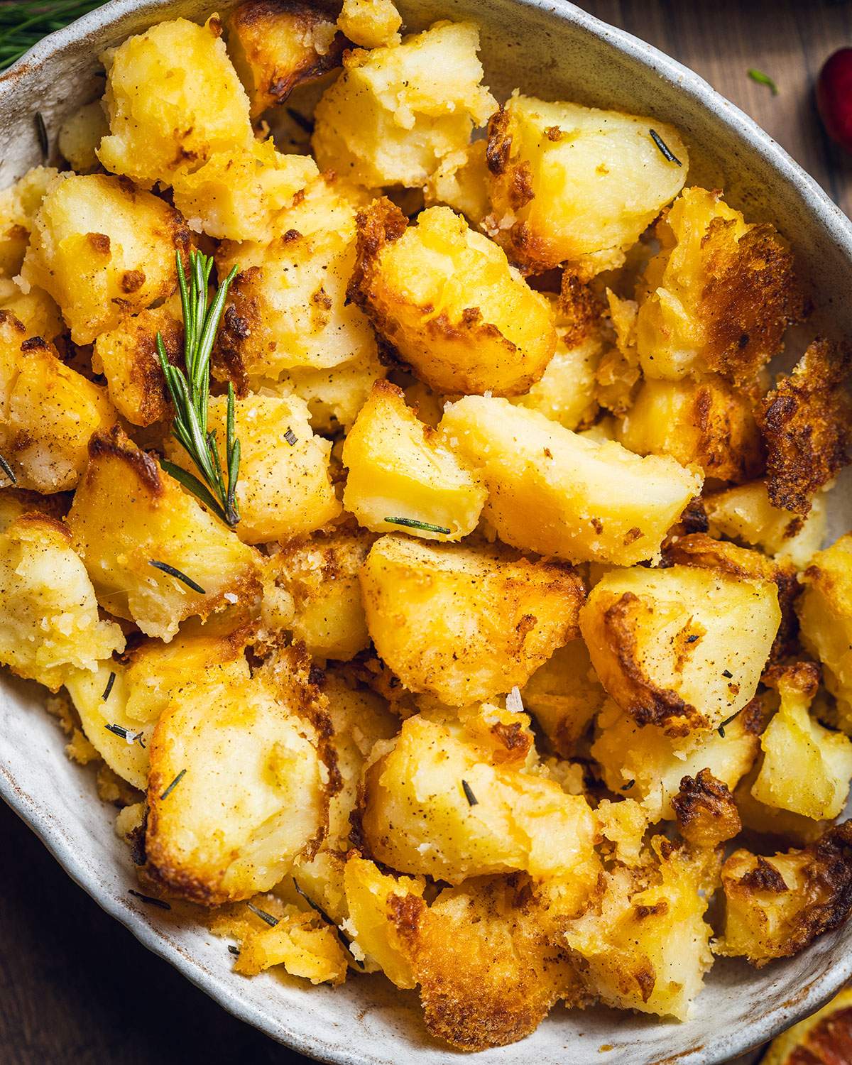 Crispy vegan roast potatoes in a serving dish up close with fresh rosemary in top.
