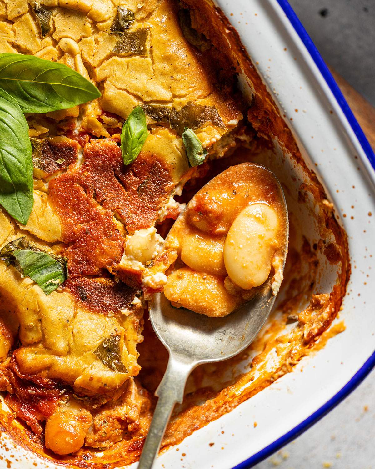 A serving spoon with cheesy butter beans in a tomato sauce on the corner of a baking dish.