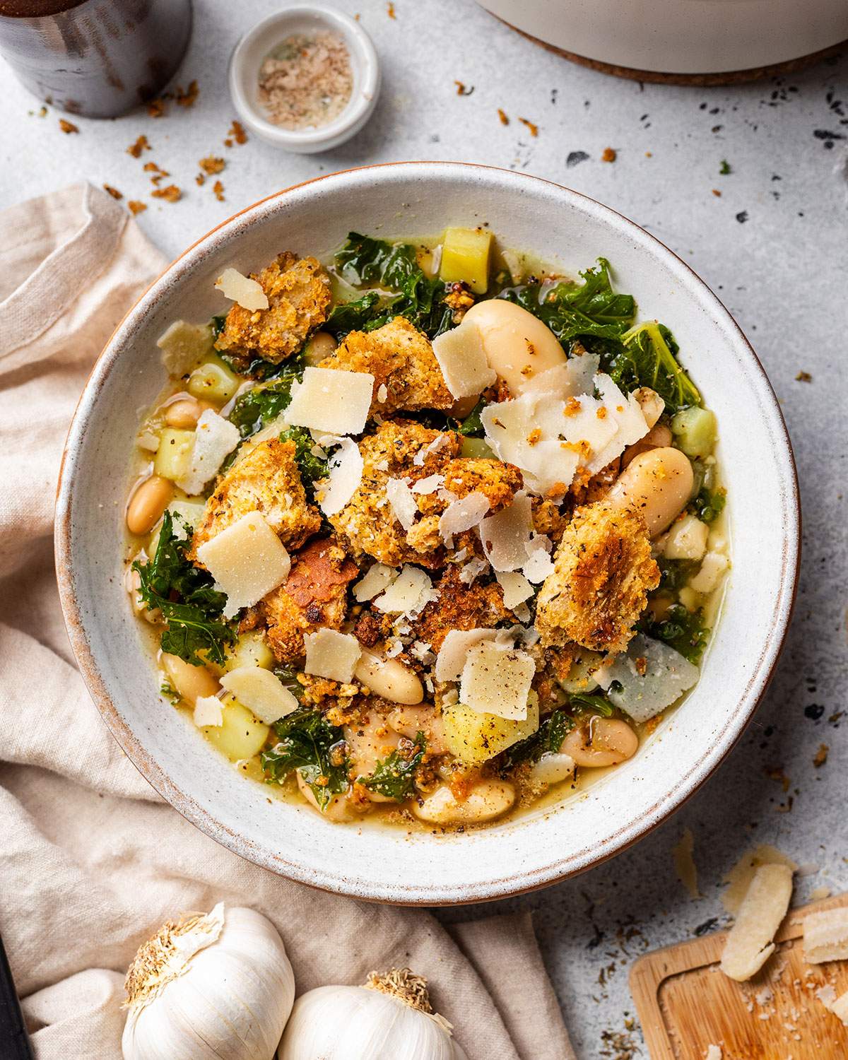 A bowl full of rustic butter bean and kale soup with kale, homemade chunky croutons and shavings of vegan parmesan.