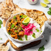 A bowl of chickpea and sweet potato curry on the dinner table, with rice, pickled onions, naan bread and fresh lime on top.