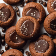 Stacked vegan espresso thumbprint cookies on a white table with espresso beans surrounding them.