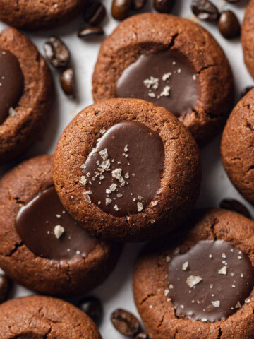 Stacked vegan espresso thumbprint cookies on a white table with espresso beans surrounding them.
