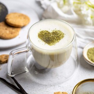 Frothy vanilla matcha latte with a matcha heart on top and cookies next to it.