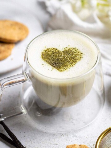 Frothy vanilla matcha latte with a matcha heart on top and cookies next to it.