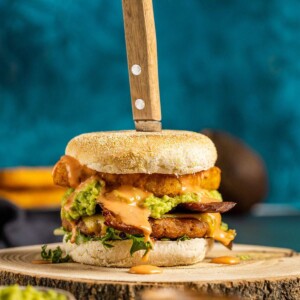 A savory vegan breakfast burger on a wooden serving tray with a knife stuck in the top of the burger.