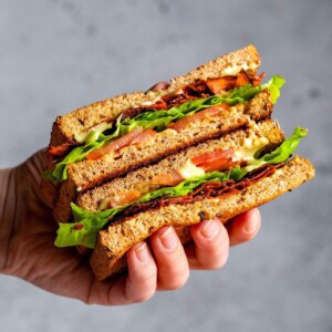 A hand holding a vegan BLT sandwich with carrot bacon.