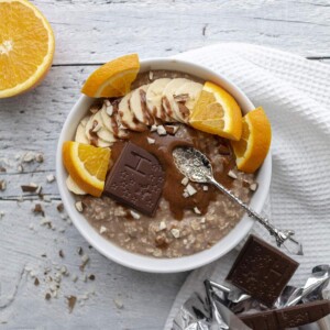 Chocolate Orange Porridgein a white bowl on a white table, topped with banana, orange slices and melted chocolate alongside nut butter.