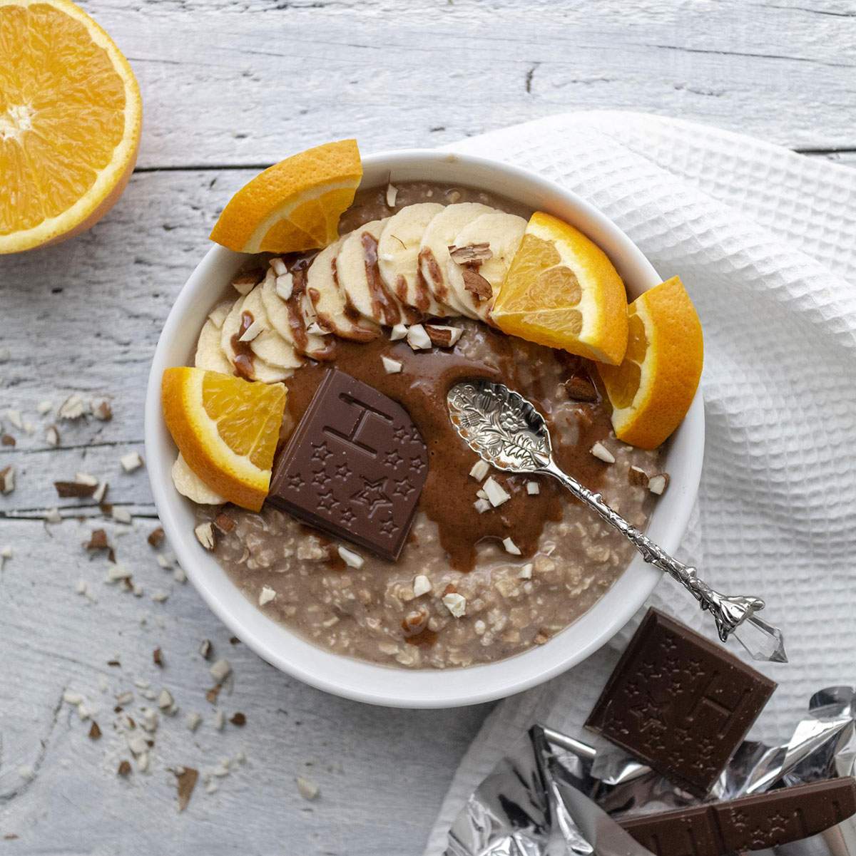 Chocolate Orange Porridgein a white bowl on a white table, topped with banana, orange slices and melted chocolate alongside nut butter.