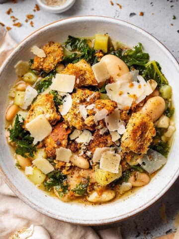A bowl full of rustic butter bean and kale soup with kale, homemade chunky croutons and shavings of vegan parmesan.