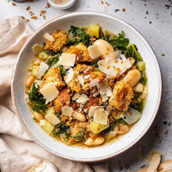 A bowl full of rustic butter bean and kale soup with kale, homemade chunky croutons and shavings of vegan parmesan.