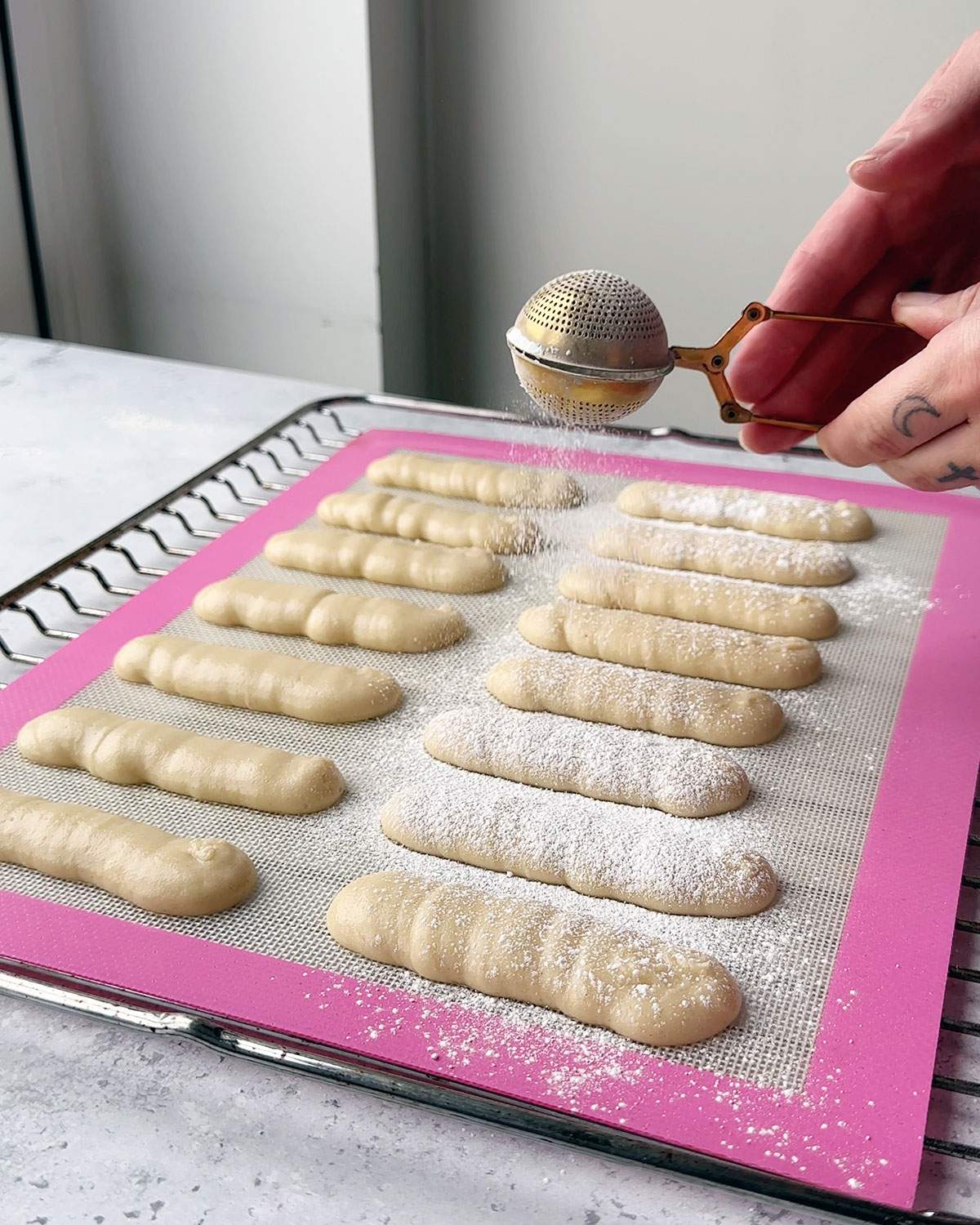 Dusting piped ladyfinger biscuit batter with icing sugar on a baking sheet.