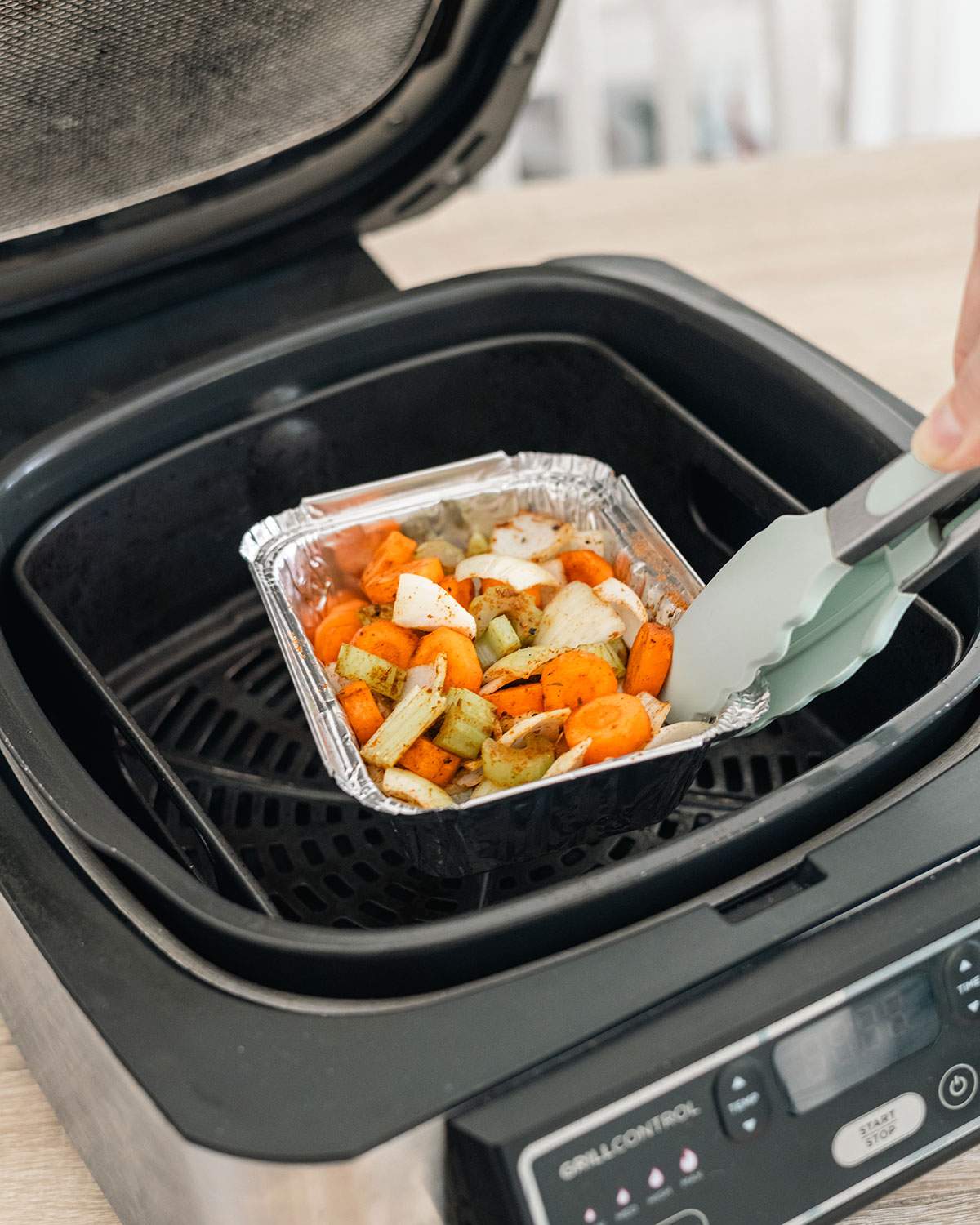Lifting an aluminum pan with cooked food out of the air fryer basket using tongs.