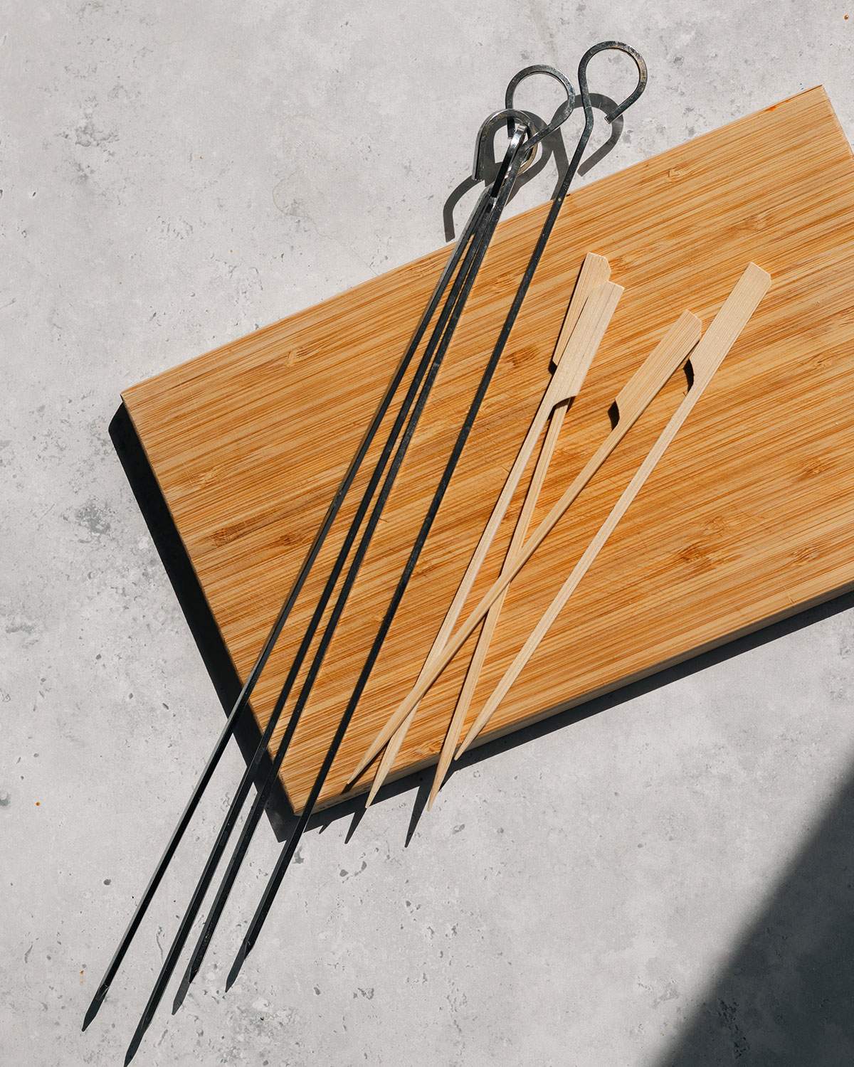 Metal skewers and wood skewers on a cutting board next to one another.