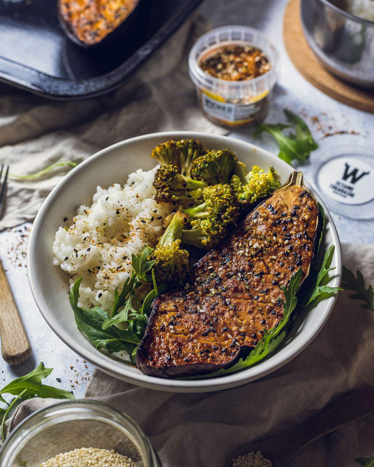 Vegan miso roasted aubergine in a white bowl served with sushi rice, rocket salad and steamed broccoli.