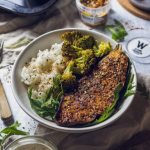 Vegan miso roasted aubergine in a white bowl served with sushi rice, rocket salad and steamed broccoli.