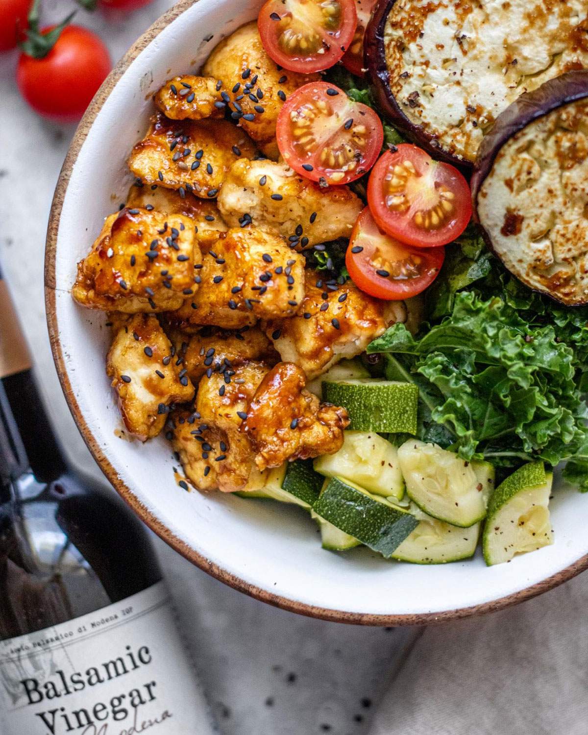 A colourful bowl filled with roasted vegetables and topped with sticky balsamic tofu is shown up close with a bottle of Balsamic Vinegar next to it.