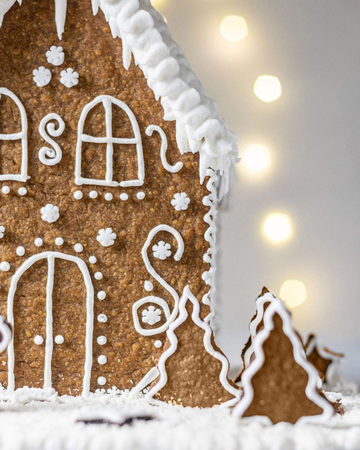 Snowy gingerbread trees decorated in front of a gingerbread house.