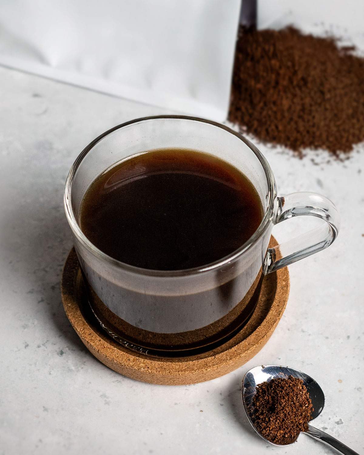 A cup of black coffee on a table next to coffee grounds and a spoon.