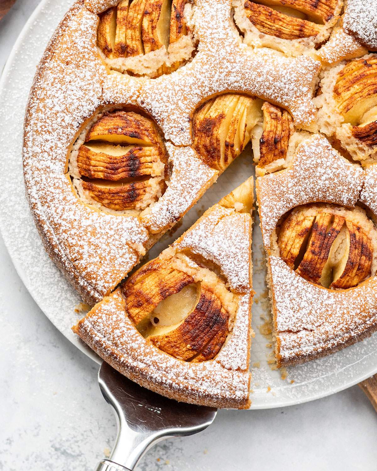 Whole vegan German apple cake dusted with icing sugar and topped with baked apples, with one slice removed and served on a white surface.