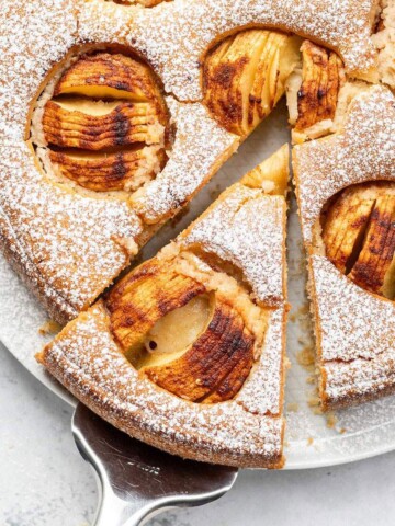 Whole vegan German apple cake dusted with icing sugar and topped with baked apples, with one slice removed and served on a white surface.