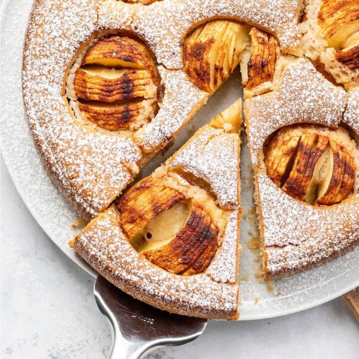 Whole vegan German apple cake dusted with icing sugar and topped with baked apples, with one slice removed and served on a white surface.