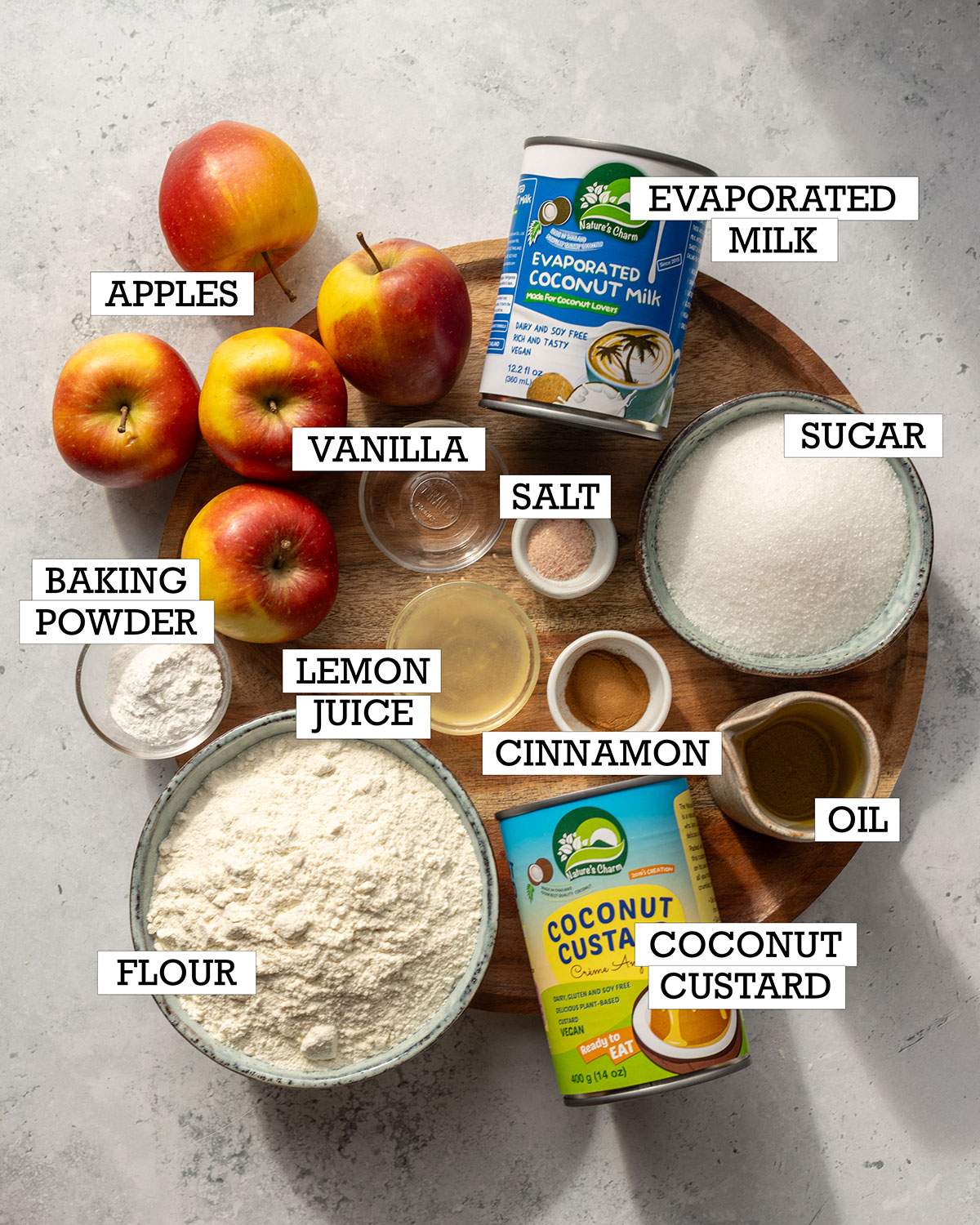 Ingredients for vegan German apple cake arranged on a wooden board, including apples, flour, sugar, coconut custard, evaporated coconut milk, oil, cinnamon, lemon juice, vanilla, baking powder, and salt.