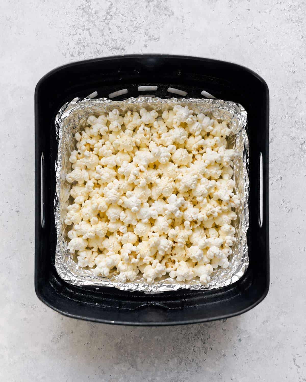 Cooked popcorn covering aluminium foil inside a black air fryer basket, viewed from above.