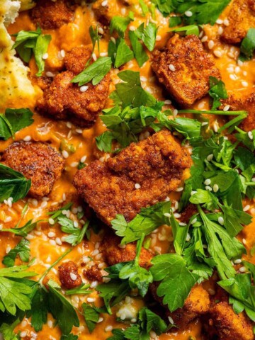 Crispy curried tofu pieces served over creamy chickpea curry, topped with fresh herbs and sesame seeds, with naan on the side.