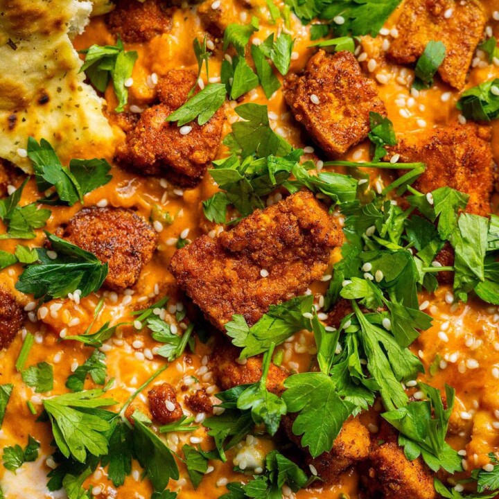 Crispy curried tofu pieces served over creamy chickpea curry, topped with fresh herbs and sesame seeds, with naan on the side.