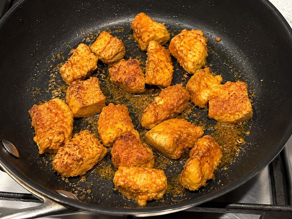 Tofu cubes coated in spices and cornstarch frying in a skillet until golden and crispy for vegan curried tofu.