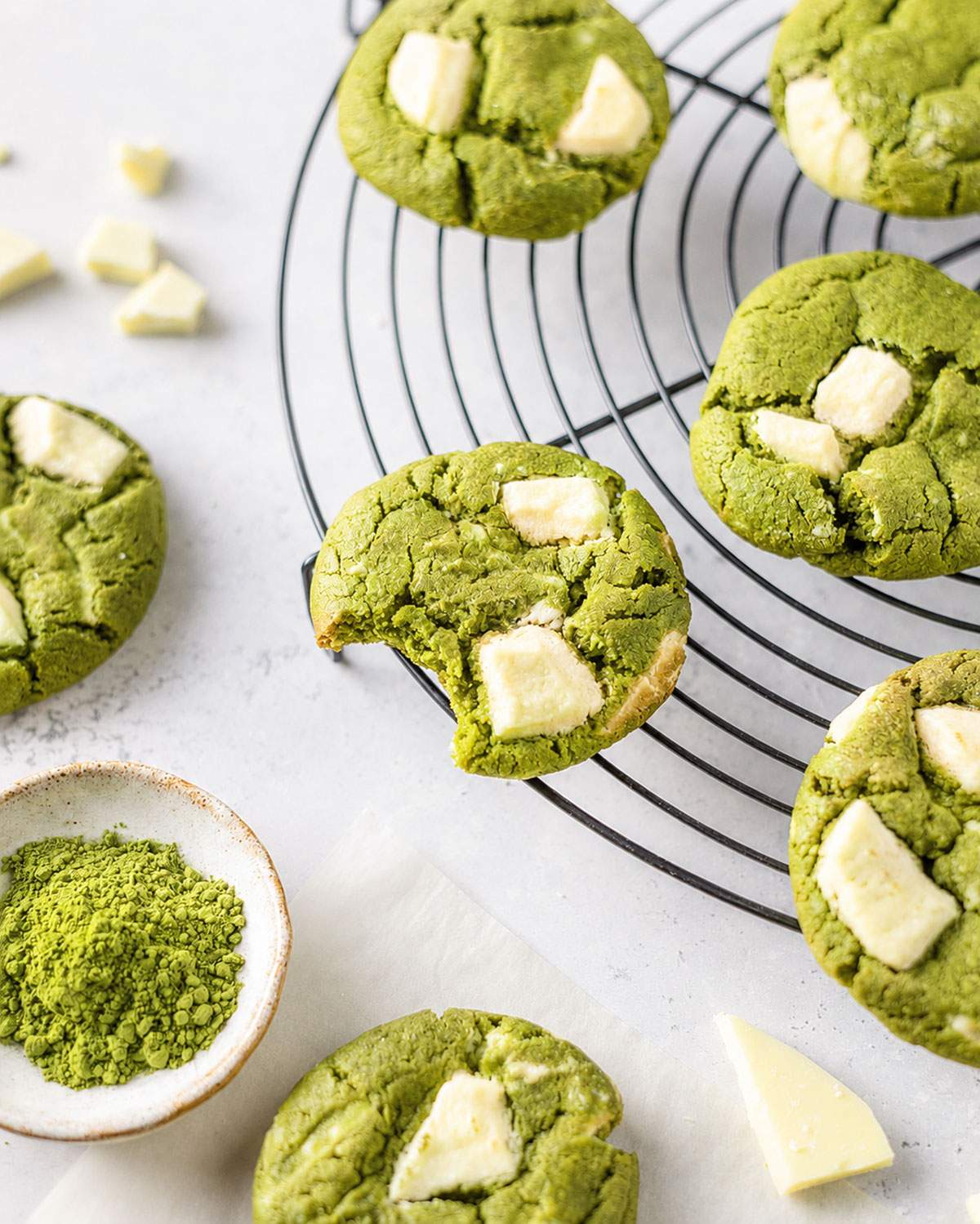 A bitten matcha cookie showing its soft centre, surrounded by white chocolate matcha cookies on a cooling rack.