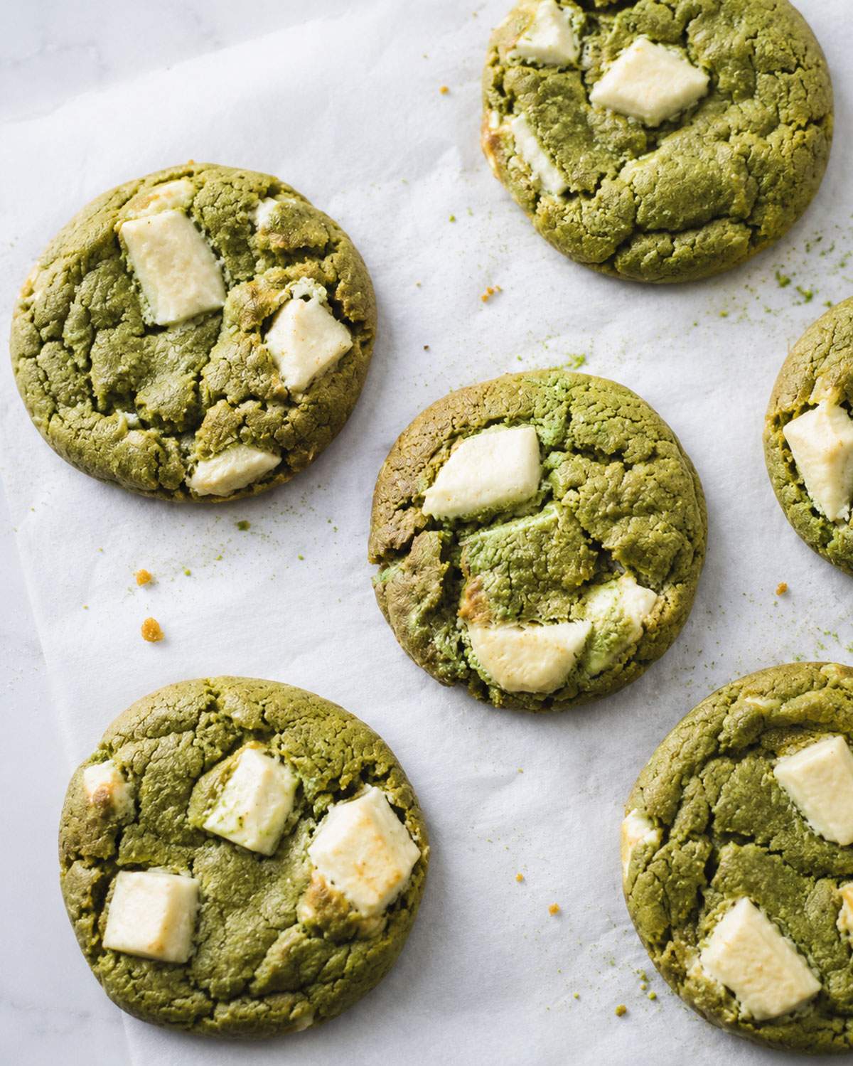 Soft and chewy matcha cookies with white chocolate chunks arranged casually on baking parchment with a few scattered crumbs.