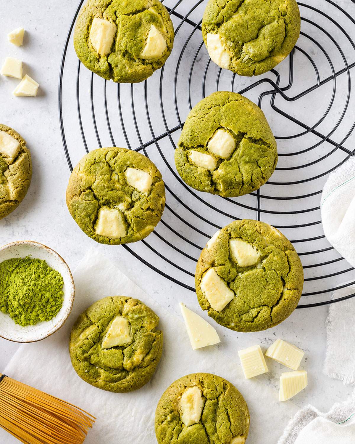 Vegan matcha white chocolate cookies cooling on a round wire rack alongside matcha powder and pieces of white chocolate.
