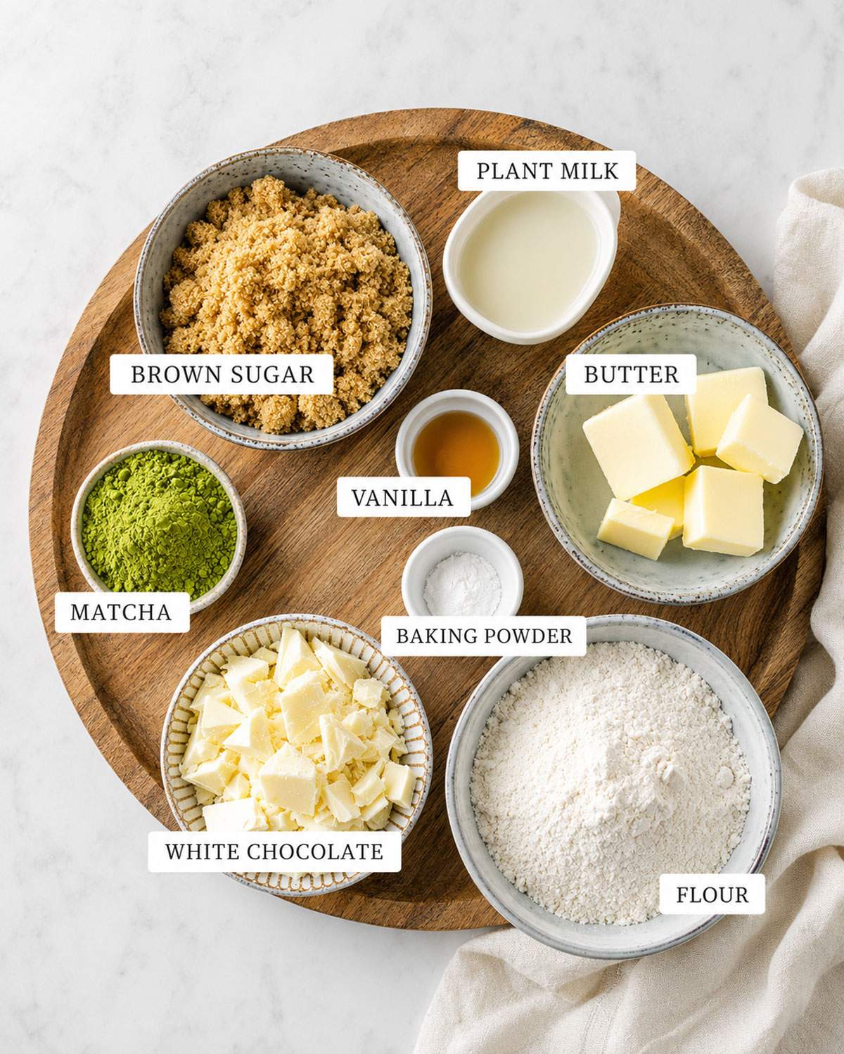 Ingredients for vegan matcha cookies arranged on a wooden board, including matcha powder, flour, brown sugar, plant milk, vegan butter and white chocolate.