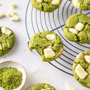 A bitten matcha cookie showing its soft centre, surrounded by white chocolate matcha cookies on a cooling rack.