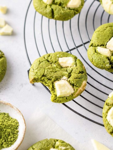 A bitten matcha cookie showing its soft centre, surrounded by white chocolate matcha cookies on a cooling rack.