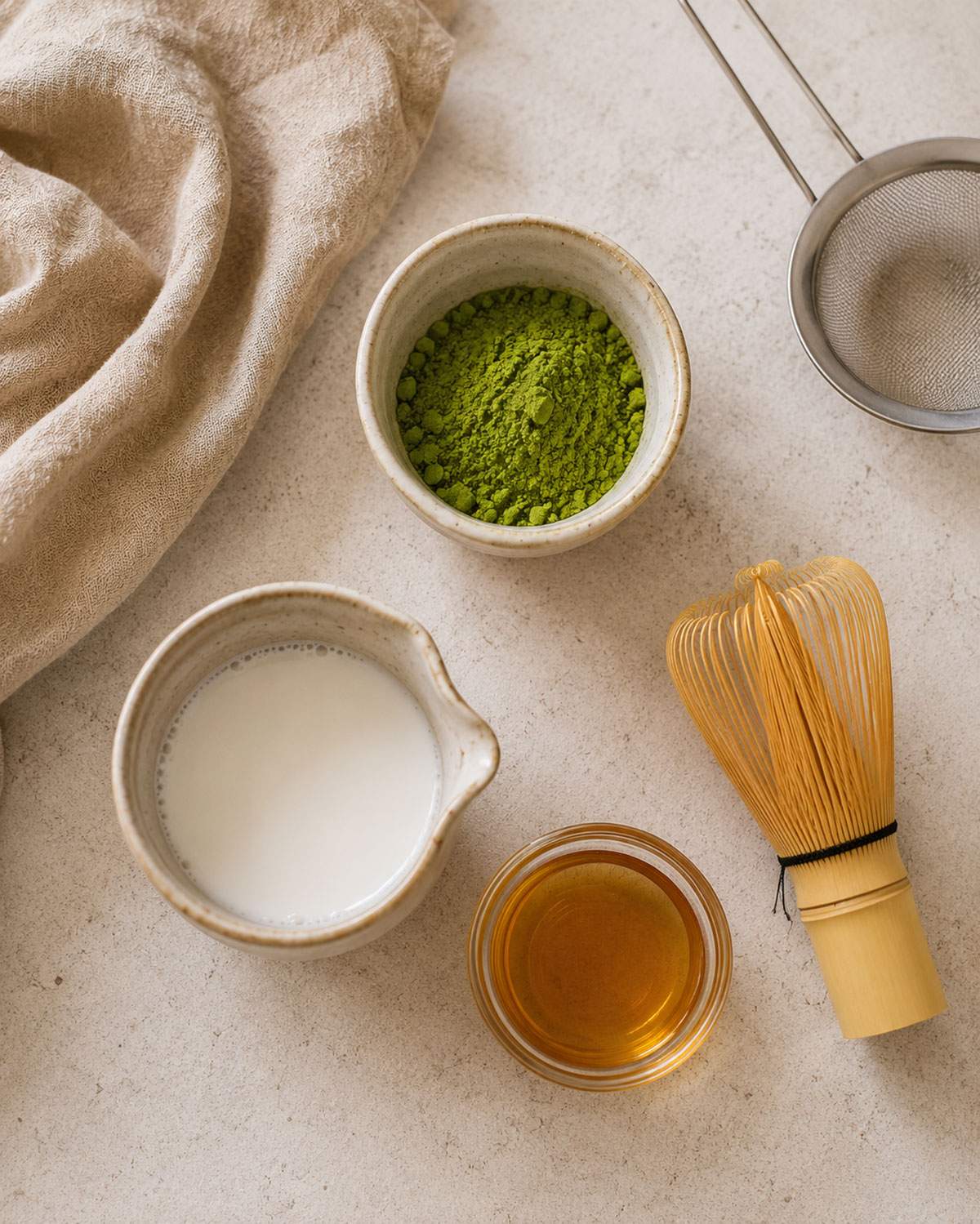Overhead view of matcha latte ingredients, including matcha powder, plant milk, maple syrup and a bamboo whisk.