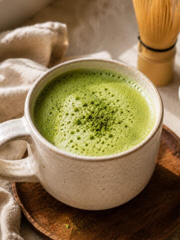 Close-up of a frothy matcha latte in a rustic ceramic mug, styled with a bamboo whisk and matcha powder.
