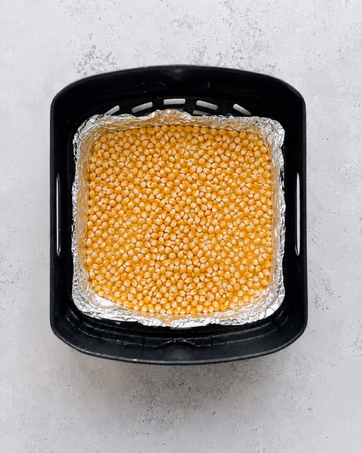 Unpopped popcorn kernels spread in a single layer over aluminium foil in a Ninja air fryer basket.