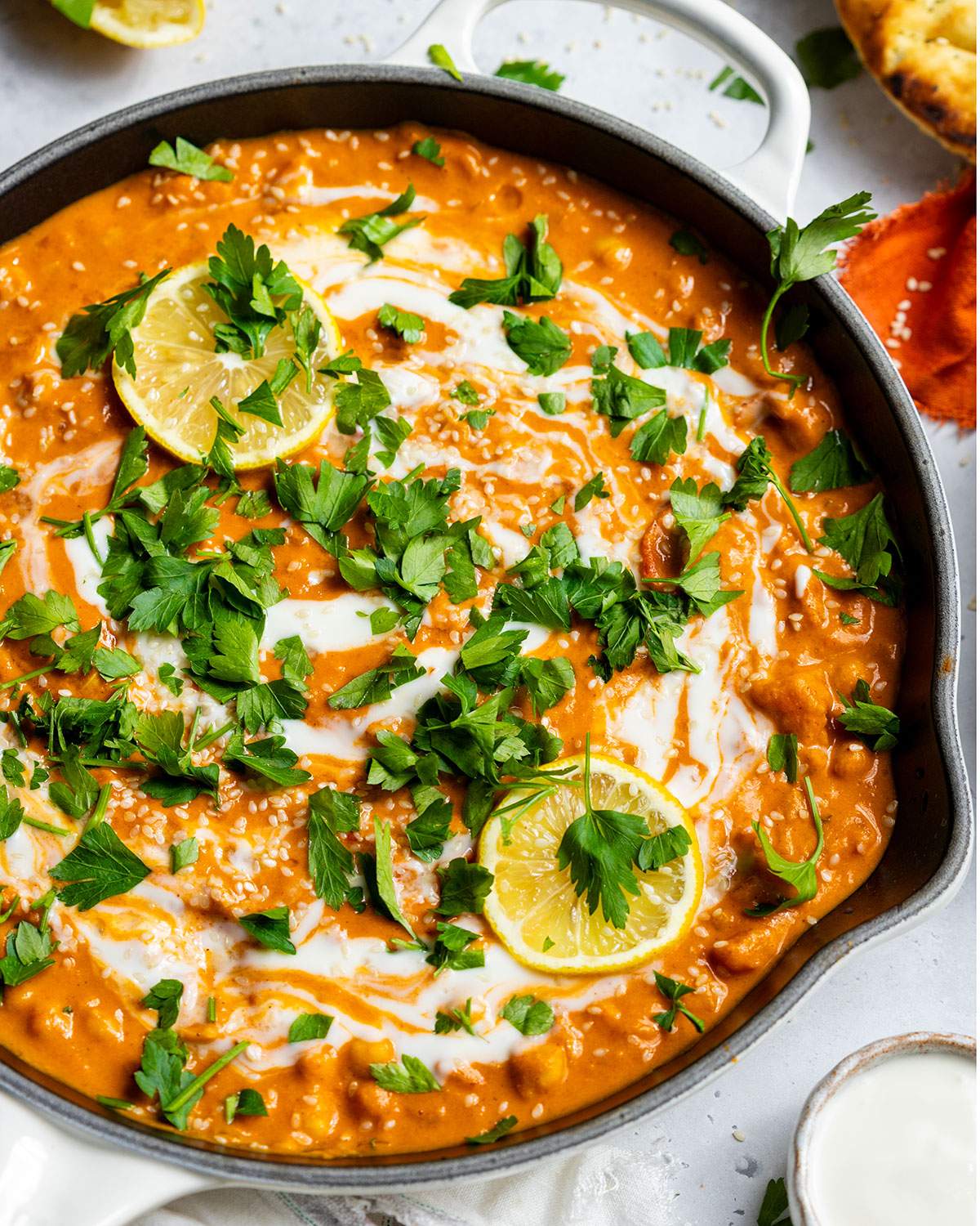 Vegan butter chickpea curry garnished with coconut cream, parsley, lemon slices, and sesame seeds, served in a white skillet with naan.