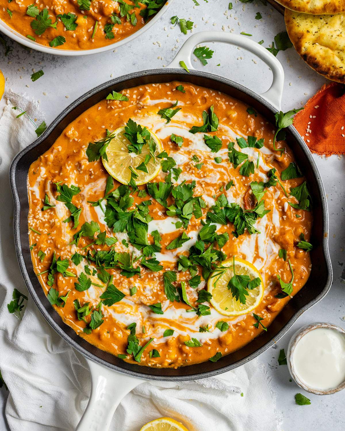 Creamy vegan butter chickpea curry in a white skillet topped with coconut cream, fresh parsley, lemon slices, and sesame seeds.