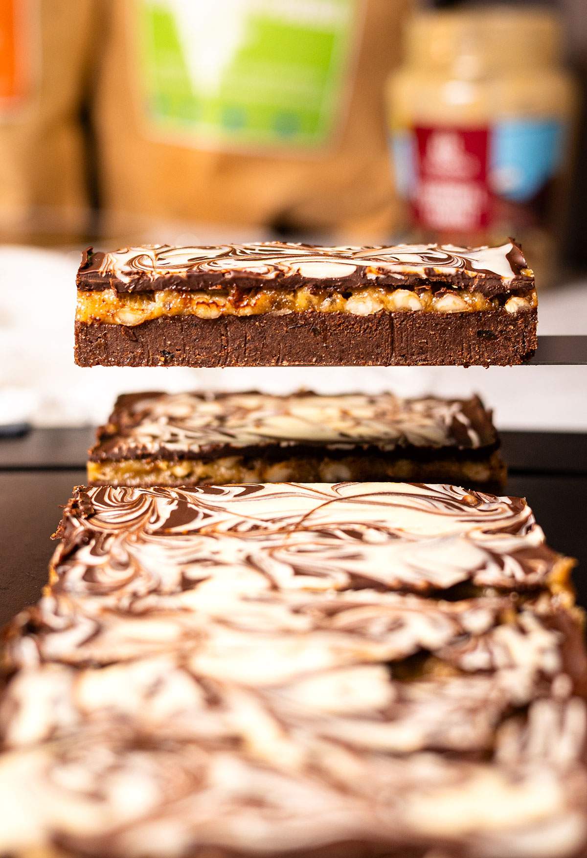 Chocolate date caramel protein bar lifted above a tray, showing its thick chocolate base, golden filling, and marbled topping.
