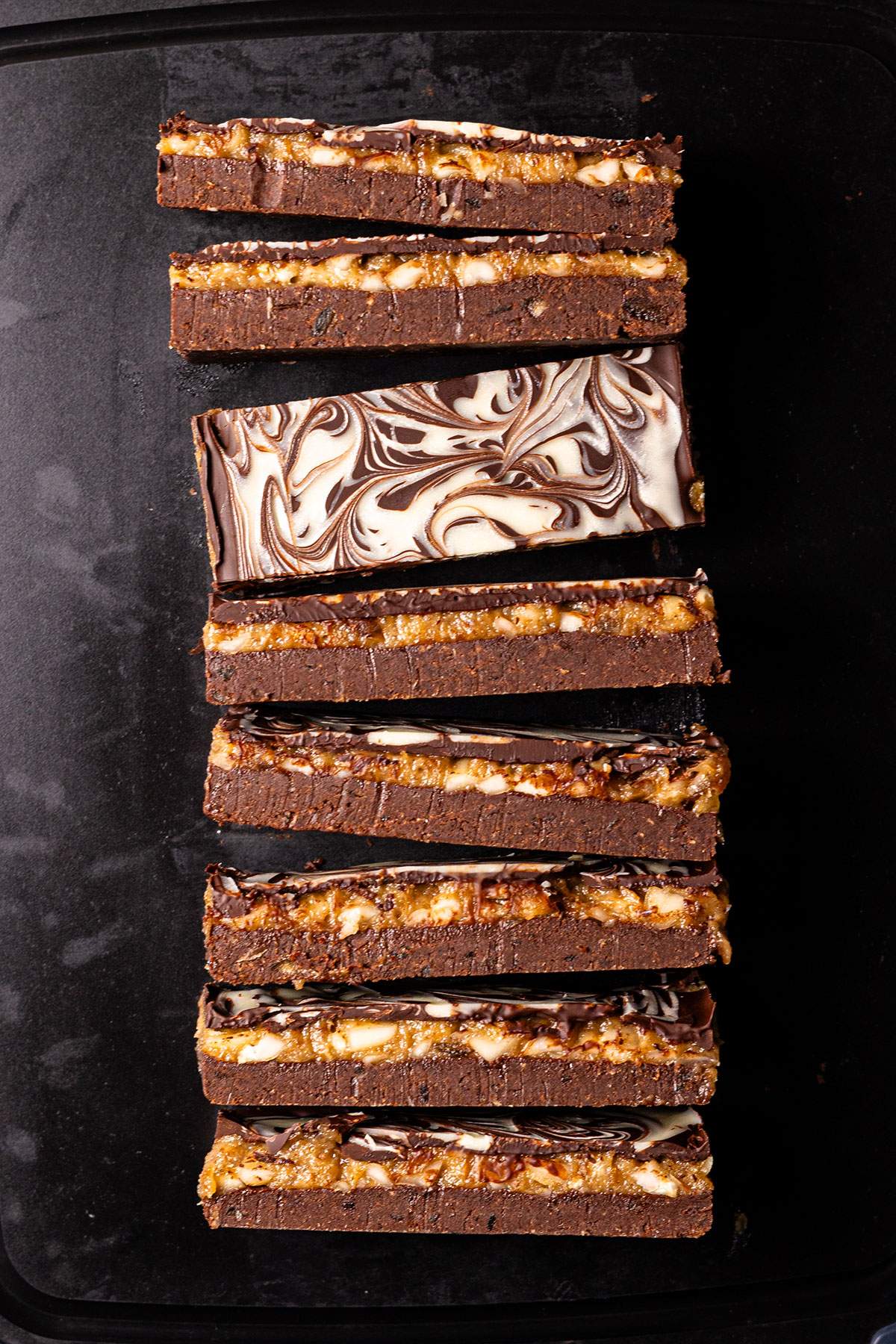 Date caramel protein bars arranged in a row on a black tray, with exposed chocolate layers and a marbled chocolate topping.