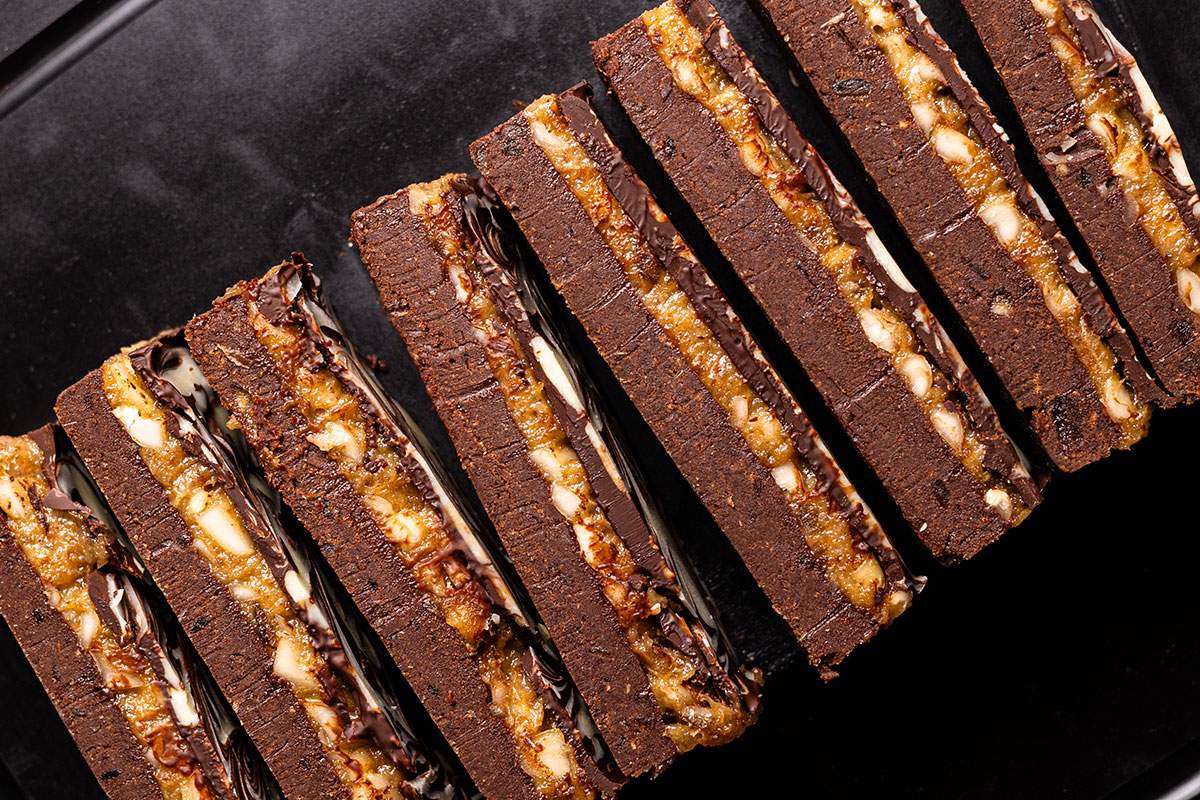 Chocolate date caramel protein bars arranged diagonally on a black tray, showing the thick base and textured caramel filling.