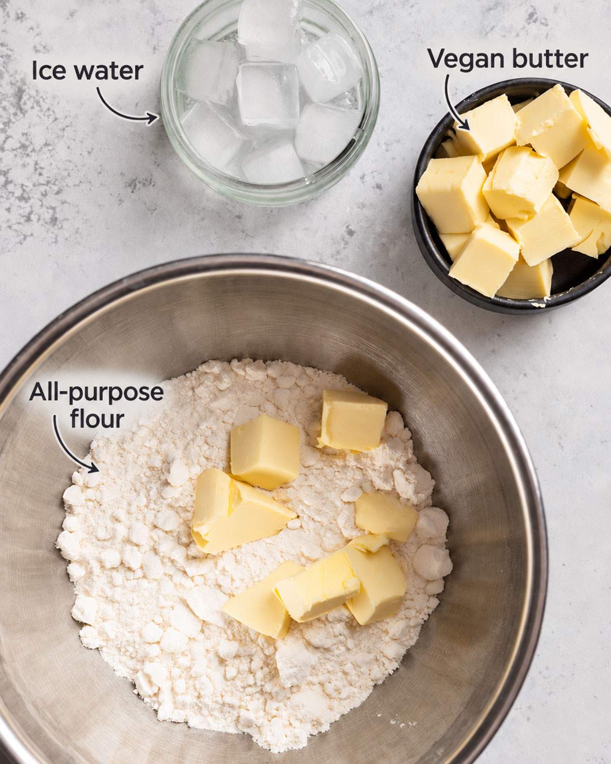 Labelled ingredients for a vegan tart base such as ice water and cold vegan butter next to a mixing bowl with flour.