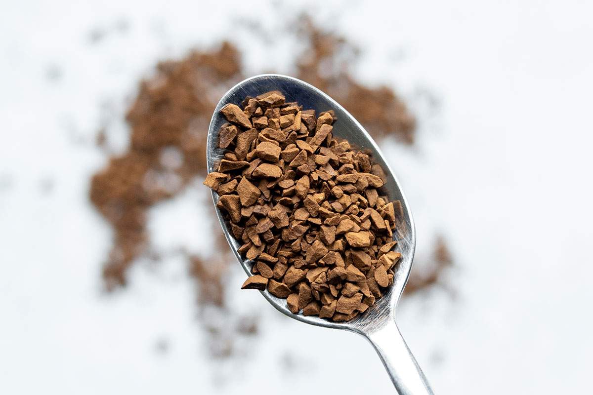 Metal teaspoon filled with instant coffee granules on a pale stone surface.